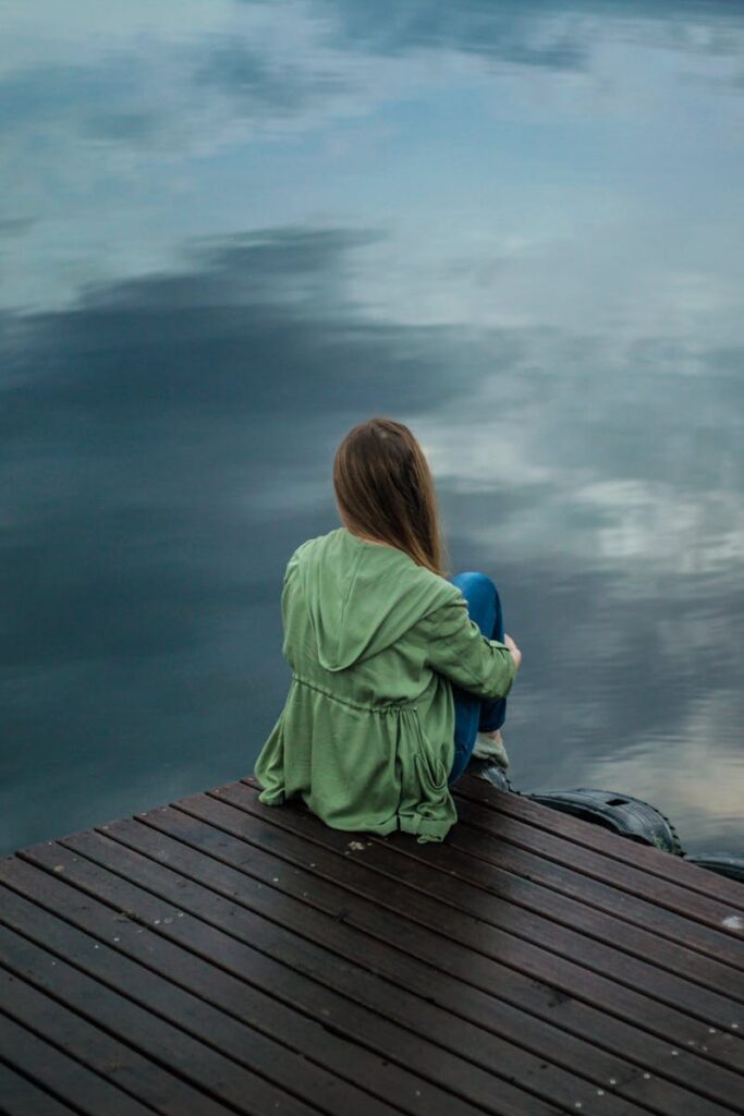 Blog A woman sits on a wooden dock, reflecting by a calm lake under a cloudy sky.