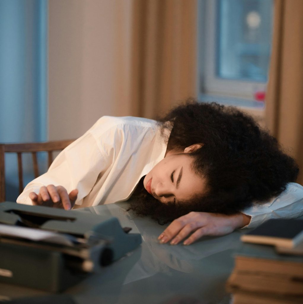 pexels photo 7970667 7970667 Woman with curly hair rests tiredly on desk near typewriter, showcasing work fatigue.